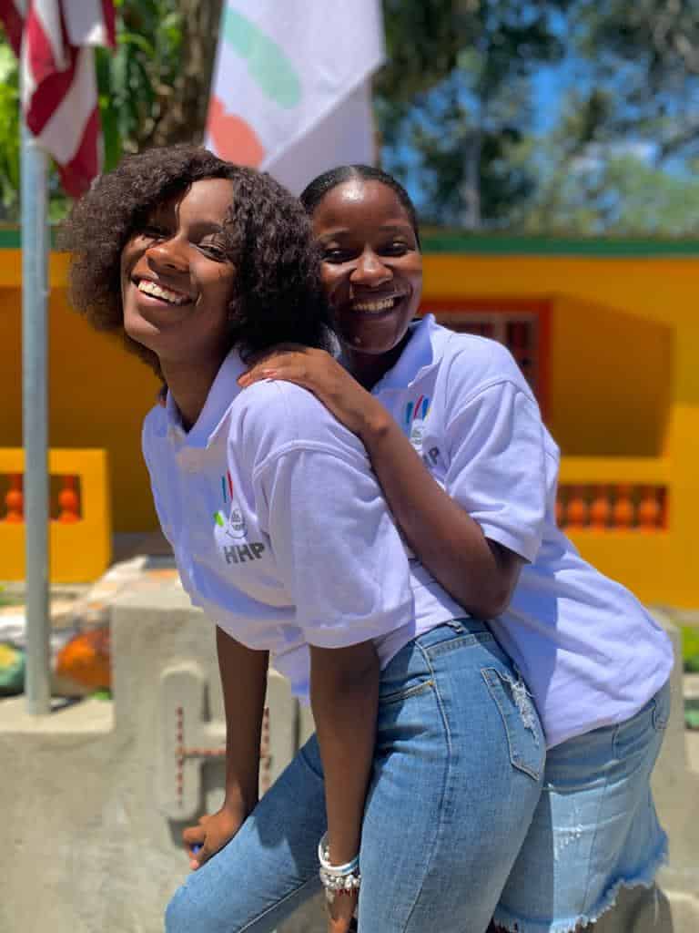 Two female students hug in uniform in front of American, Happy Haitian Education and Happy Haitian Productions flags