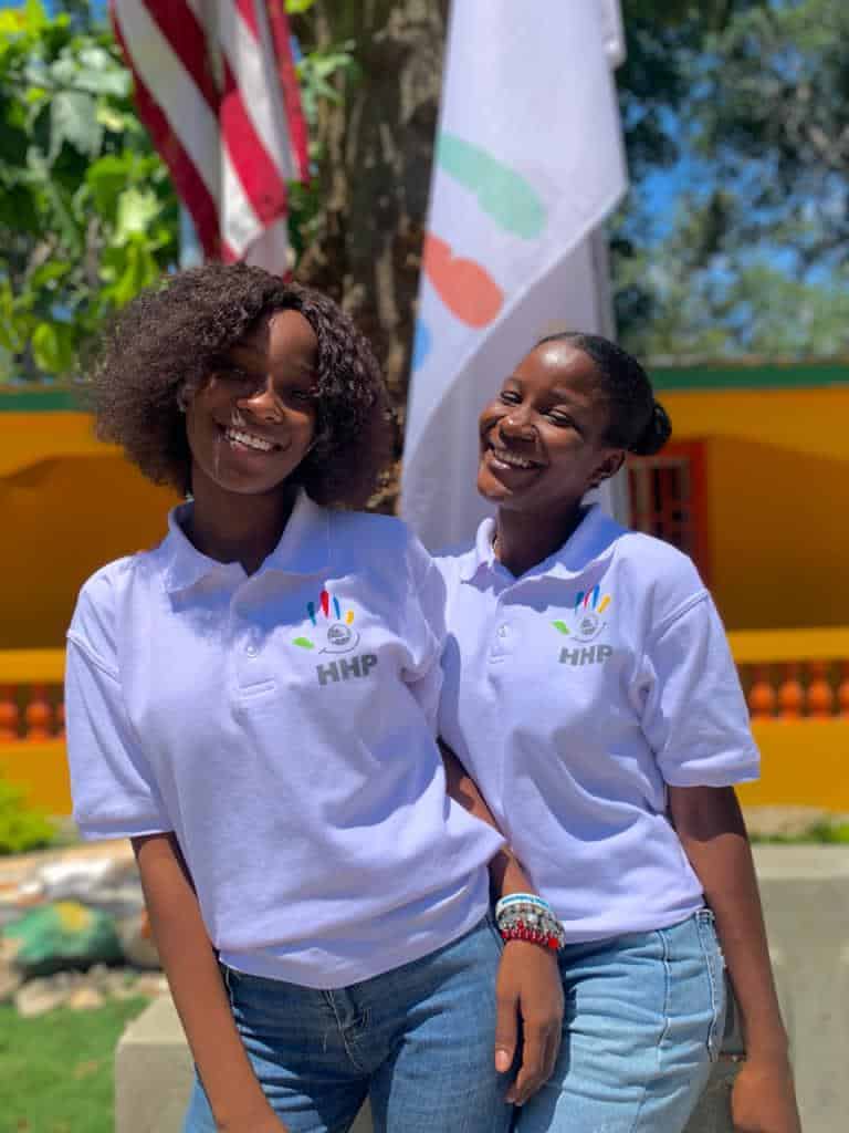 Two female students smile in uniform in front of American, Happy Haitian Education and Happy Haitian Productions flags